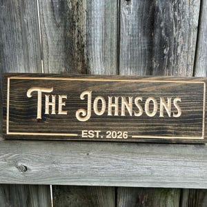 May include: A rectangular wooden sign with the engraved text "The Johnsons" and "EST. 2026" in a light color against a dark stained background. The sign has a framed border.