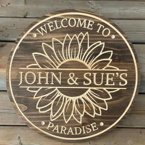 May include: A round, wooden sign with a sunflower design and the words "WELCOME TO JOHN & SUE'S PARADISE" carved into it. The sign is stained a dark brown with a lighter wood sunflower design.