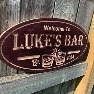 May include: Oval wooden sign with the text "Welcome to LUKE'S BAR" in a carved, light-colored font. The sign also features the text "Est. 2024" and two etched glasses. The sign is a dark brown color.