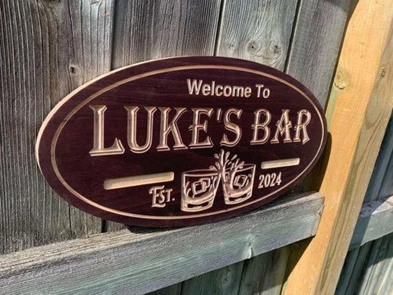 May include: Oval wooden sign with the text "Welcome to LUKE'S BAR" in a carved, light-colored font. The sign also features the text "Est. 2024" and two etched glasses. The sign is a dark brown color.
