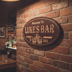 May include: Oval wooden sign with the text "Welcome To LUKE'S BAR" in a decorative font. The sign includes the establishment date "Est. 2024" and two etched glasses. The sign is mounted on a brick wall.