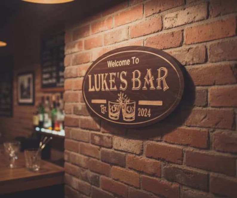 May include: Oval wooden sign with the text "Welcome To LUKE'S BAR" in a decorative font. The sign includes the establishment date "Est. 2024" and two etched glasses. The sign is mounted on a brick wall.
