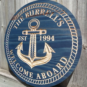 May include: A round, navy blue wooden sign with a carved anchor design. The sign includes the text "THE BURRELL'S WELCOME ABOARD EST 1994" in a nautical style, with a rope border. The anchor is a light wood color.