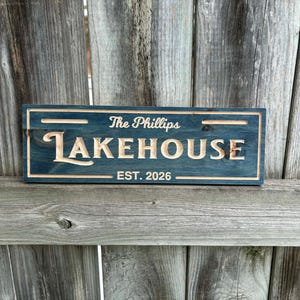 May include: A rectangular wooden sign with the words "The Phillips LAKEHOUSE EST. 2026" carved into it. The sign is painted a dark teal color with the lettering in a lighter, natural wood tone. The sign is displayed against a weathered wooden background.