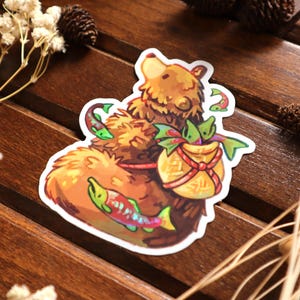 Op de afbeelding: Een sticker met een cartoonbeer met bruine vacht, die een gouden tas draagt die met een rood lint is vastgebonden. De tas is gevuld met groene en rode vissen. Een kleurrijke vis staat op de voorgrond.