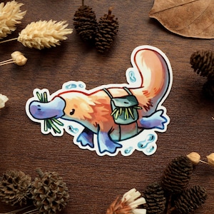 Peut inclure: Un autocollant coloré représentant un ornithorynque de dessin animé avec un sac à dos, tenant de l'herbe dans son bec. L'ornithorynque est orange, bleu et blanc. L'autocollant a une bordure blanche et est entouré d'éléments décoratifs.