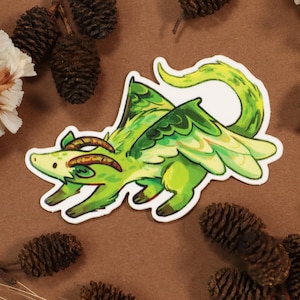 May include: A die-cut sticker of a green and yellow fantasy creature with horns and wings. The sticker has a white border and is set against a brown background with pinecones and dried flowers.