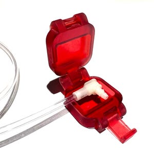 Puede incluir: Un dispositivo médico de plástico rojo con una tapa con bisagras, abierto para revelar un conector de plástico blanco. Un tubo transparente se extiende desde el dispositivo. El dispositivo está sobre un fondo blanco.