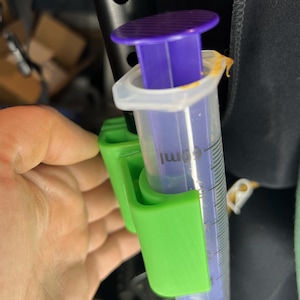 May include: A clear 60ml syringe with purple plunger and cap, held in a bright green holder. The syringe has black measurement markings. The holder is attached to a black surface.