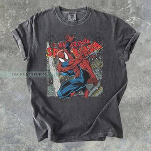 May include: Dark gray t-shirt featuring a vintage-style graphic of Spider-Man in a dynamic pose. The design includes the words "AMAZING SPIDER-MAN" in red and yellow. The shirt has rolled-up sleeves and a classic crew neck.