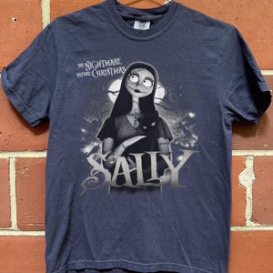 May include: Navy blue t-shirt featuring Sally from "The Nightmare Before Christmas." The graphic shows Sally holding a black cat, with the movie title above and her name below in silver lettering. The shirt is hanging on a metal hanger.