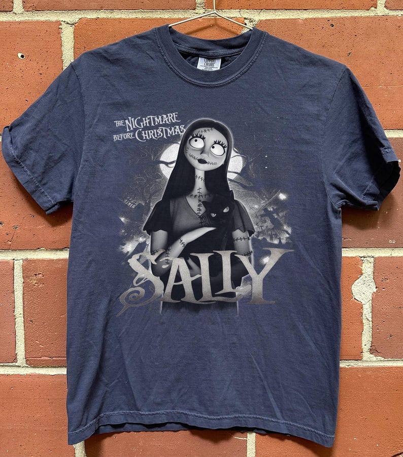May include: Navy blue t-shirt featuring Sally from "The Nightmare Before Christmas." The graphic shows Sally holding a black cat, with the movie title above and her name below in silver lettering. The shirt is hanging on a metal hanger.