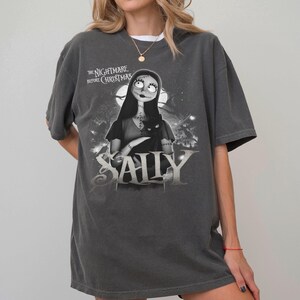 May include: Dark gray t-shirt featuring a graphic of Sally from "The Nightmare Before Christmas." The design includes Sally holding a black cat, with the word "SALLY" in large, stylized letters. The shirt has short sleeves and a crew neck.