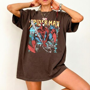 May include: Brown oversized t-shirt featuring a graphic of Spider-Man in a dynamic pose, with the text "SPIDER-MAN" in yellow. The Marvel logo is visible above the graphic. The shirt has short sleeves and a relaxed fit.