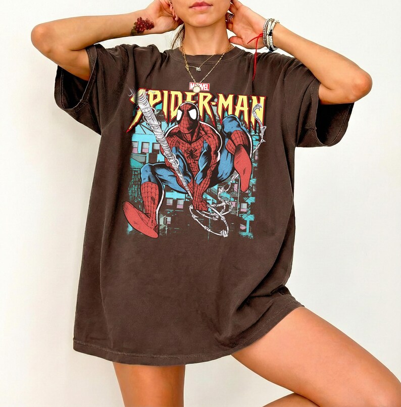 May include: Brown oversized t-shirt featuring a graphic of Spider-Man in a dynamic pose, with the text "SPIDER-MAN" in yellow. The Marvel logo is visible above the graphic. The shirt has short sleeves and a relaxed fit.