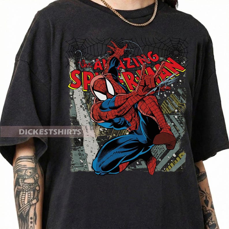 May include: Dark gray t-shirt featuring a comic book illustration of Spider-Man. The graphic shows Spider-Man in a dynamic pose with the words "AMAZING SPIDER-MAN" in red and yellow. The shirt has the text "DICKESTSHIRTS" on the bottom left.