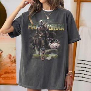 May include: Dark gray t-shirt featuring a graphic of The Mandalorian and Grogu from Star Wars. The design includes the text "STAR WARS" above the title "THE MANDALORIAN" and the phrase "THIS IS THE WAY" below the image.