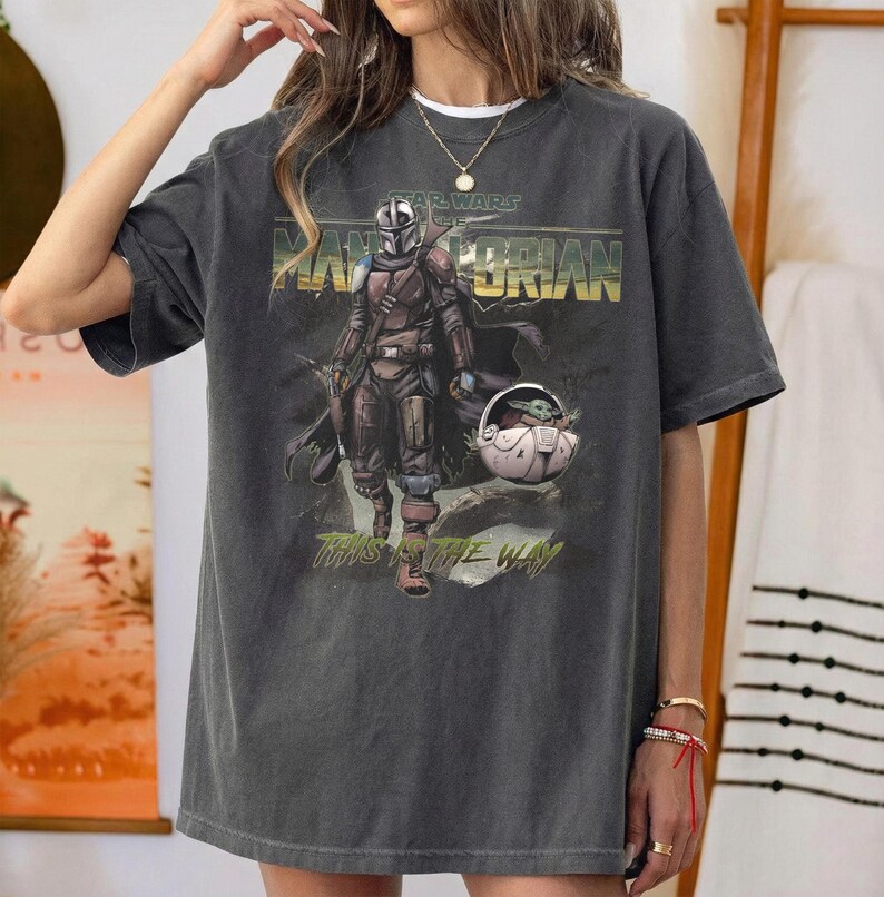 May include: Dark gray t-shirt featuring a graphic of The Mandalorian and Grogu from Star Wars. The design includes the text "STAR WARS" above the title "THE MANDALORIAN" and the phrase "THIS IS THE WAY" below the image.