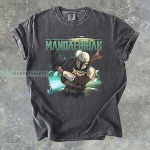 May include: Dark gray t-shirt featuring a graphic of The Mandalorian holding Grogu, with the word "MANDALORIAN" in green. The shirt has rolled-up sleeves and a vintage look.