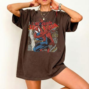 May include: Brown t-shirt featuring a graphic of Spider-Man in a dynamic pose. The design includes the words "AMAZING SPIDER-MAN" in red and yellow. The shirt has short sleeves and a relaxed fit.