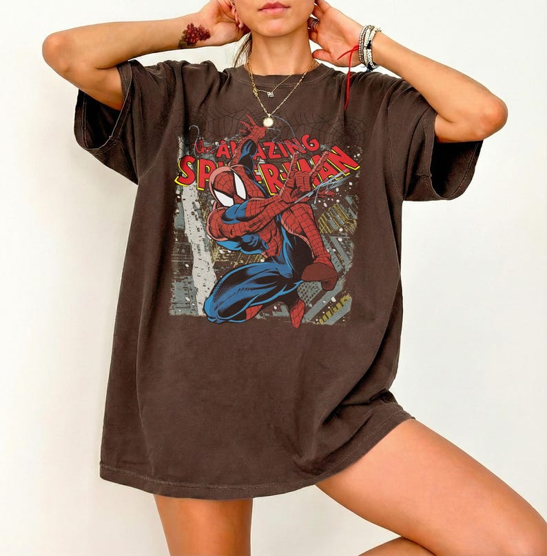 May include: Brown t-shirt featuring a graphic of Spider-Man in a dynamic pose. The design includes the words "AMAZING SPIDER-MAN" in red and yellow. The shirt has short sleeves and a relaxed fit.