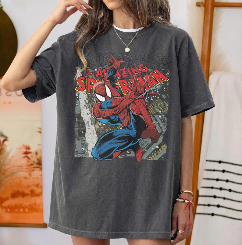 May include: Dark gray t-shirt featuring a vintage comic book graphic of Spider-Man. The design includes the words "AMAZING SPIDER-MAN" in red and yellow. The superhero is depicted in his classic red and blue suit, with a cityscape background.