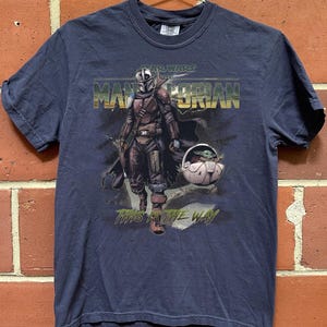 May include: Navy blue t-shirt featuring a graphic of The Mandalorian and Grogu from Star Wars. The text "STAR WARS THE MANDALORIAN" and "THIS IS THE WAY" are printed on the front. The shirt is hanging on a metal hanger.