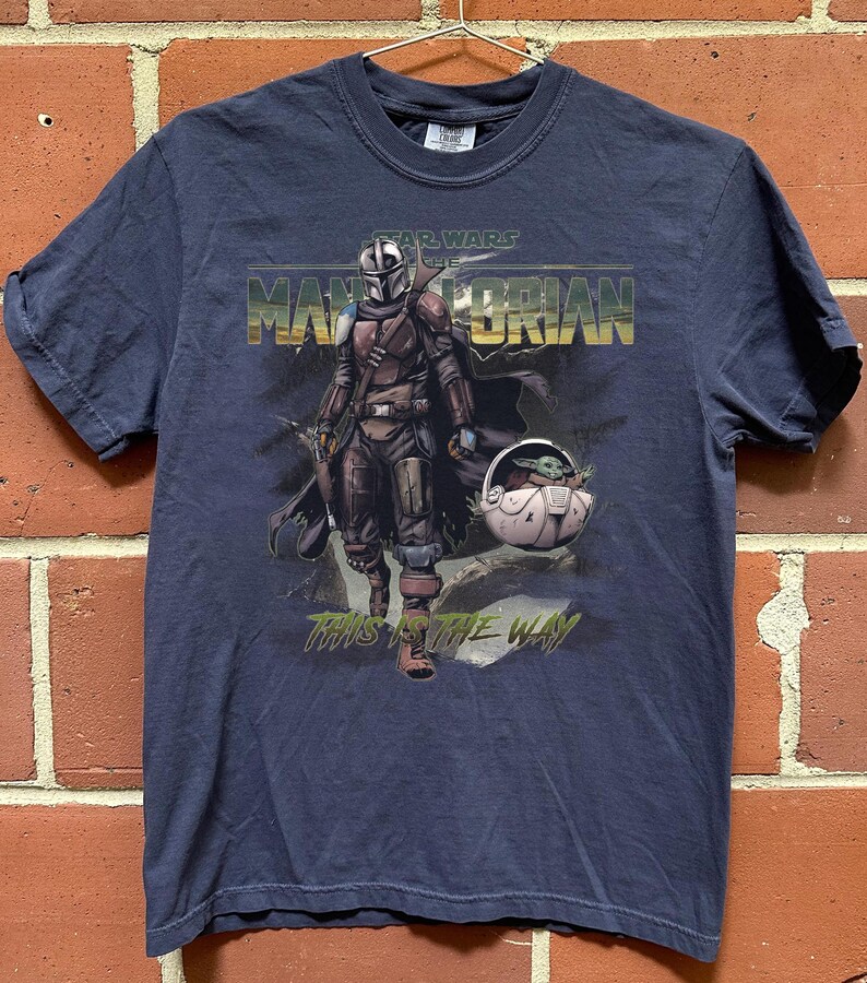 May include: Navy blue t-shirt featuring a graphic of The Mandalorian and Grogu from Star Wars. The text "STAR WARS THE MANDALORIAN" and "THIS IS THE WAY" are printed on the front. The shirt is hanging on a metal hanger.