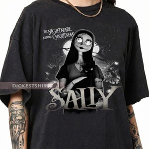 May include: Dark gray t-shirt featuring Sally from "The Nightmare Before Christmas." The design includes Sally holding a black cat, with the movie title above and her name in large letters below. The shirt has a distressed look.