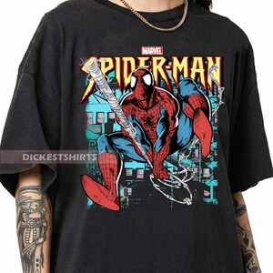 May include: Dark gray t-shirt featuring a Spider-Man graphic. The design includes the Marvel logo and the words "Spider-Man" in red and yellow. The illustration shows Spider-Man in a dynamic pose with his web.