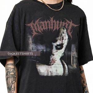 Puede incluir: Camiseta negra con un estampado gráfico de un rostro distorsionado y un paisaje urbano. La palabra "Manhunt" está impresa en una fuente gótica sobre el rostro. El texto "DICKESTSHIRTS" está impreso en la parte inferior izquierda de la camiseta.