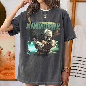May include: Dark gray t-shirt featuring a graphic of The Mandalorian holding Grogu, with the text "MANDALORIAN" in green. The design is set against a futuristic, neon-lit background. The shirt has a vintage, worn look.