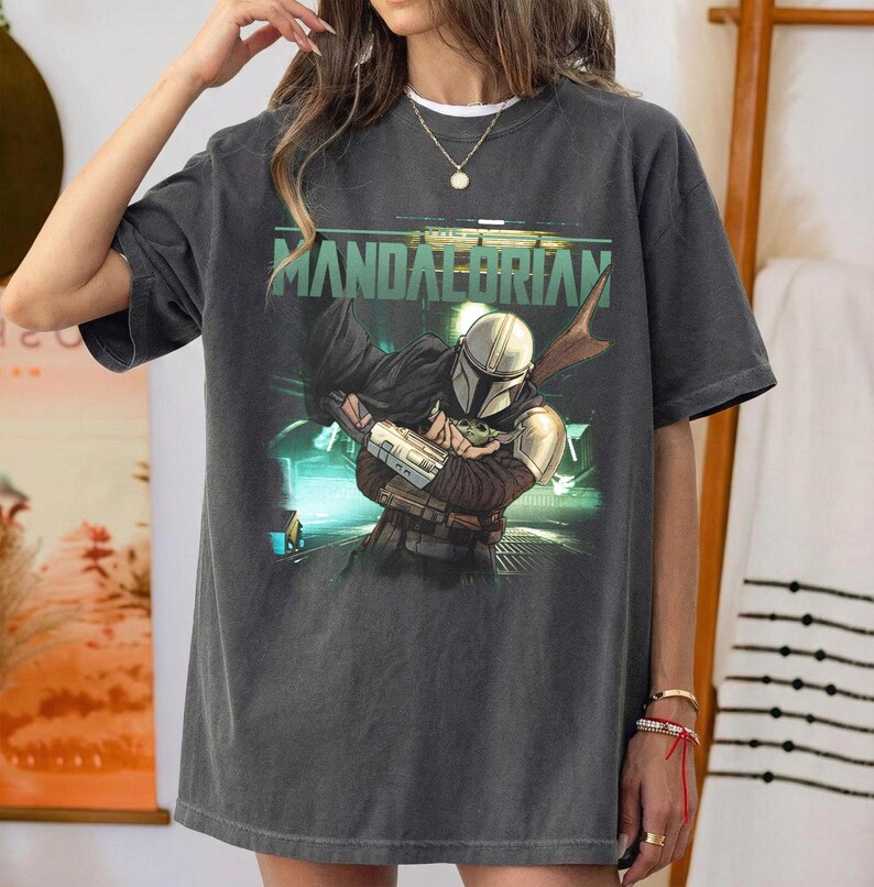 May include: Dark gray t-shirt featuring a graphic of The Mandalorian holding Grogu, with the text "MANDALORIAN" in green. The design is set against a futuristic, neon-lit background. The shirt has a vintage, worn look.