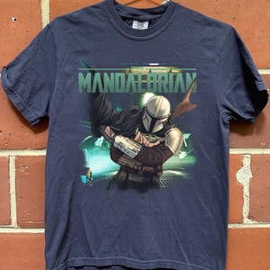 May include: Navy blue t-shirt featuring a graphic of The Mandalorian holding Grogu, with the text "MANDALORIAN" in green. The design includes a futuristic background. The shirt is hanging on a metal hanger.