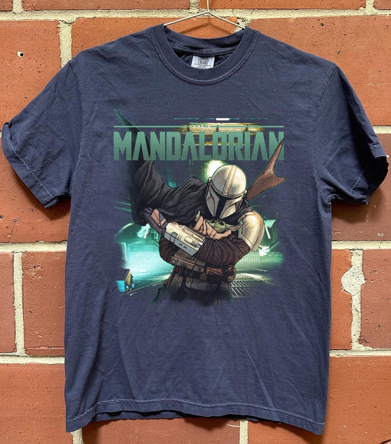 May include: Navy blue t-shirt featuring a graphic of The Mandalorian holding Grogu, with the text "MANDALORIAN" in green. The design includes a futuristic background. The shirt is hanging on a metal hanger.
