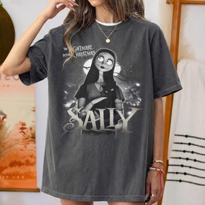 May include: Dark gray t-shirt featuring Sally from "The Nightmare Before Christmas." The graphic shows Sally holding a black cat with a moonlit background. The word "SALLY" is prominently displayed below the character.