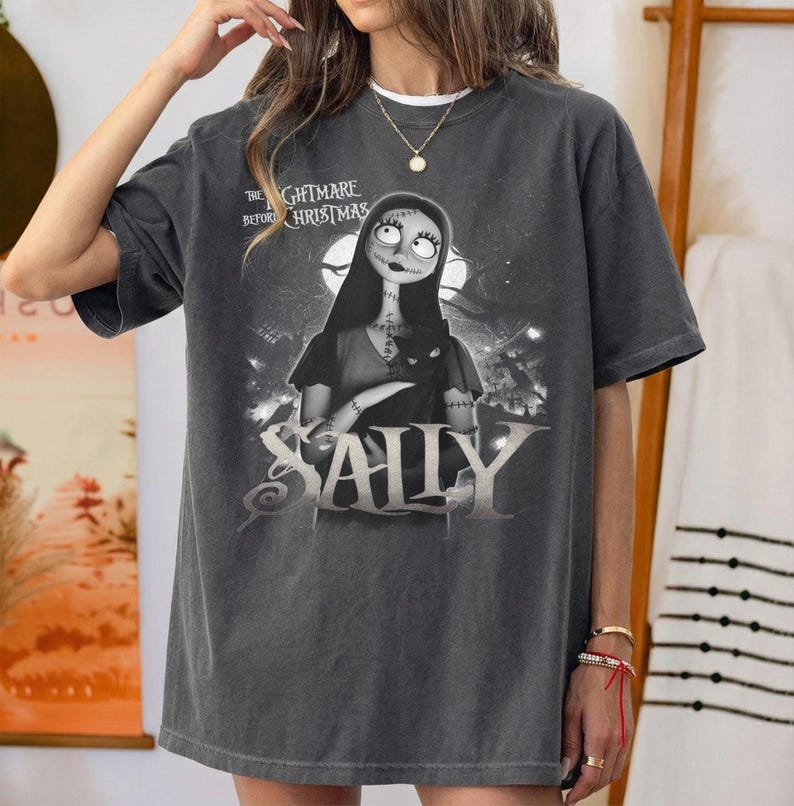 May include: Dark gray t-shirt featuring Sally from "The Nightmare Before Christmas." The graphic shows Sally holding a black cat with a moonlit background. The word "SALLY" is prominently displayed below the character.