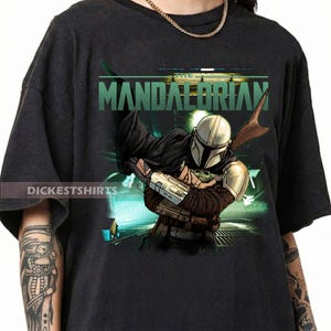 May include: Black t-shirt featuring a graphic of The Mandalorian holding Grogu, with the word "Mandalorian" in green. The design includes a helmeted figure and a small green character. The shirt has the text "Dickestshirts".