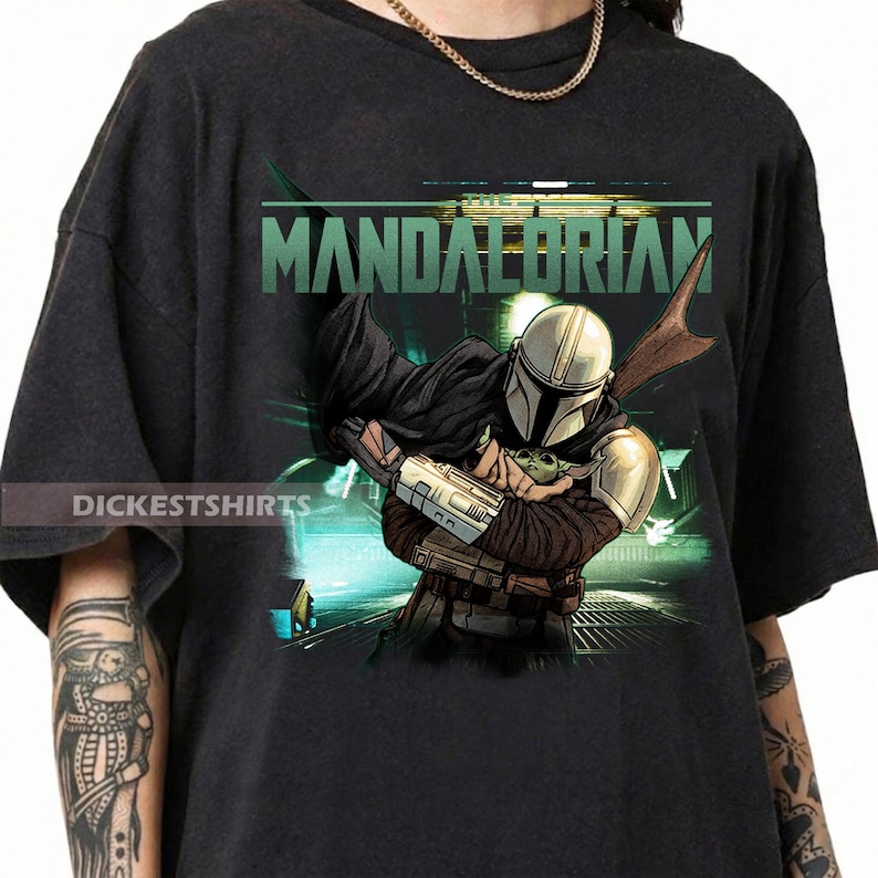 May include: Black t-shirt featuring a graphic of The Mandalorian holding Grogu, with the word "Mandalorian" in green. The design includes a helmeted figure and a small green character. The shirt has the text "Dickestshirts".