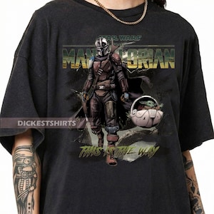 May include: Black t-shirt featuring a graphic of The Mandalorian and Grogu from Star Wars. The design includes the text "STAR WARS THE MANDALORIAN" and "THIS IS THE WAY". The image shows the Mandalorian in armor and Grogu in a carrier.