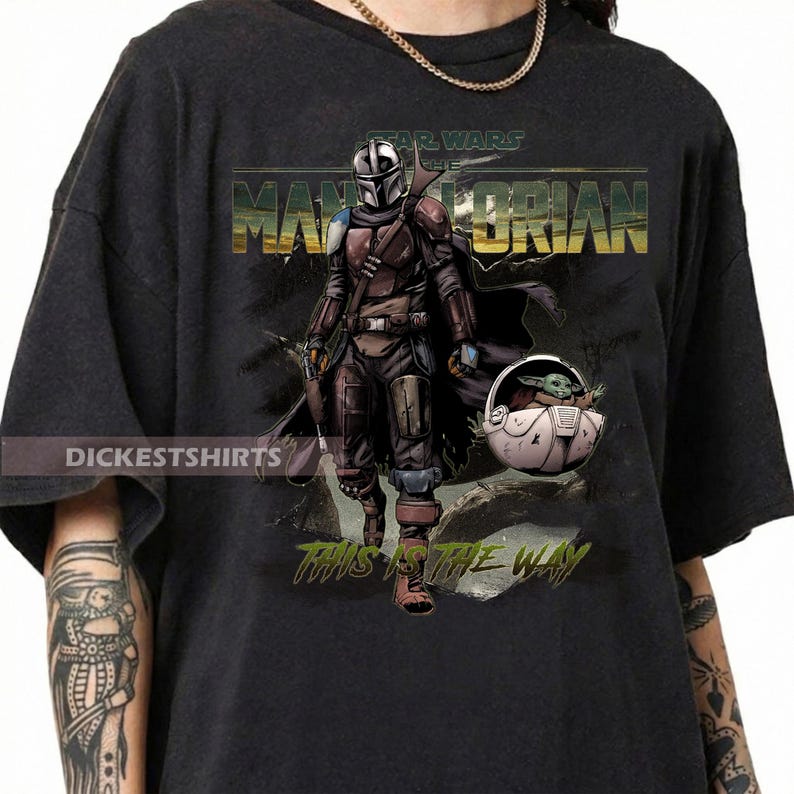 May include: Black t-shirt featuring a graphic of The Mandalorian and Grogu from Star Wars. The design includes the text "STAR WARS THE MANDALORIAN" and "THIS IS THE WAY". The image shows the Mandalorian in armor and Grogu in a carrier.