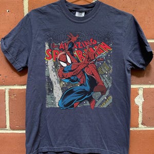 May include: Navy blue t-shirt featuring a graphic of Spider-Man in action, with the words "AMAZING SPIDER-MAN" in red and yellow. The superhero is depicted in his classic red and blue suit, with a cityscape background.