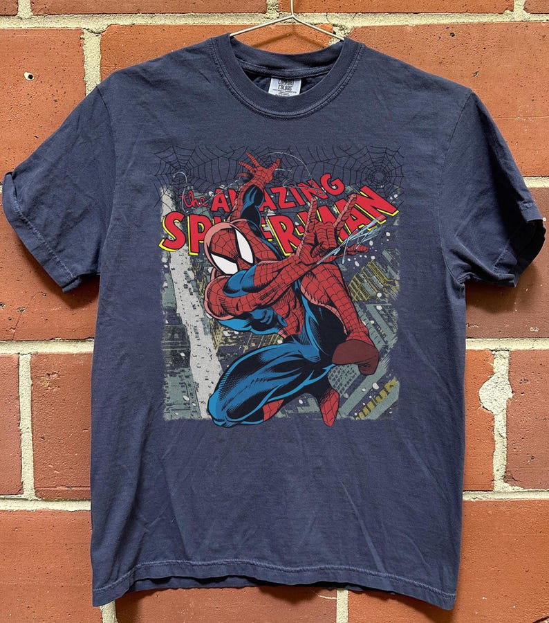 May include: Navy blue t-shirt featuring a graphic of Spider-Man in action, with the words "AMAZING SPIDER-MAN" in red and yellow. The superhero is depicted in his classic red and blue suit, with a cityscape background.