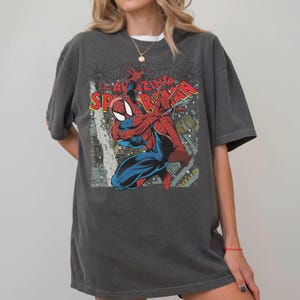 May include: Dark gray t-shirt featuring a graphic of Spider-Man in a dynamic pose, with the words "AMAZING SPIDER-MAN" in red and yellow. The design includes a cityscape and spiderwebs.