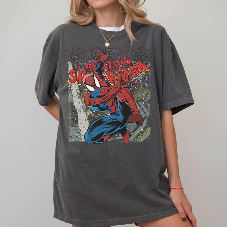 May include: Dark gray t-shirt featuring a graphic of Spider-Man in a dynamic pose, with the words "AMAZING SPIDER-MAN" in red and yellow. The design includes a cityscape and spiderwebs.