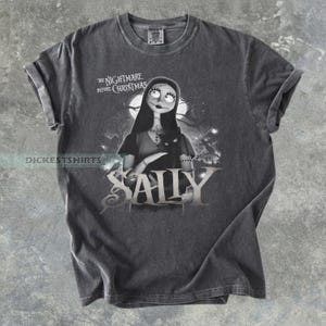 May include: Dark gray t-shirt featuring Sally from "The Nightmare Before Christmas." The graphic shows Sally with a black cat, the movie title, and the character's name. The shirt has rolled-up sleeves and a crew neck.