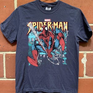 May include: Navy blue Spider-Man t-shirt featuring a graphic of the superhero in action, swinging from a web. The Marvel logo is above the Spider-Man text. The design includes red, blue, and white colors.