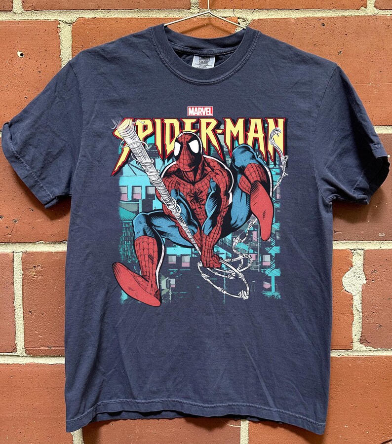 May include: Navy blue Spider-Man t-shirt featuring a graphic of the superhero in action, swinging from a web. The Marvel logo is above the Spider-Man text. The design includes red, blue, and white colors.
