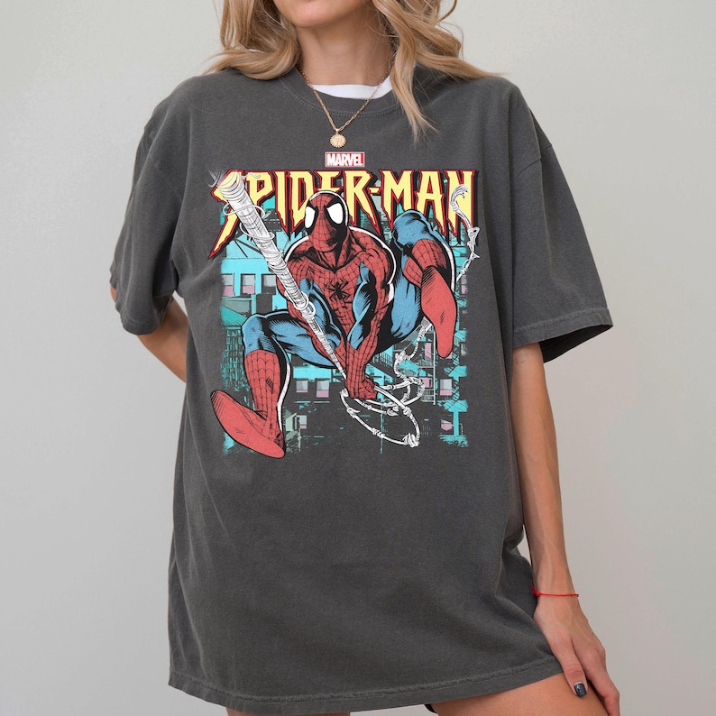 May include: Dark gray t-shirt featuring a colorful Spider-Man graphic. The design includes the Marvel logo and the words "Spider-Man" in a comic book style. The superhero is depicted in his classic red and blue suit.