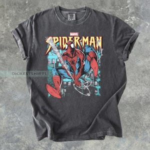 May include: Dark gray t-shirt featuring a graphic of Spider-Man in a dynamic pose, swinging from a web. The design includes the Marvel logo and the words "Spider-Man" in a bold, colorful font. The shirt has rolled-up sleeves.